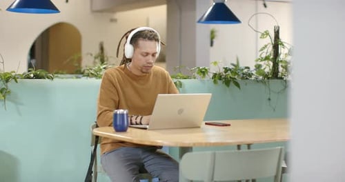 Homme afro-américain portant un pull moutarde à la table de travail, tapant, riant d'un ordinateur portable via un casque