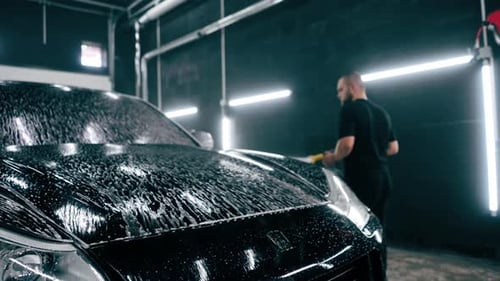 A male car wash employee applies car wash detergent to a black luxury car using spray gun in the car