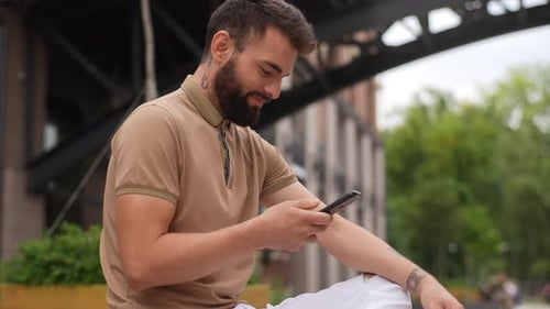 Side Lowangle View of Cheerful Young Man Sitting on Bench Outdoors Using Typing Smartphone