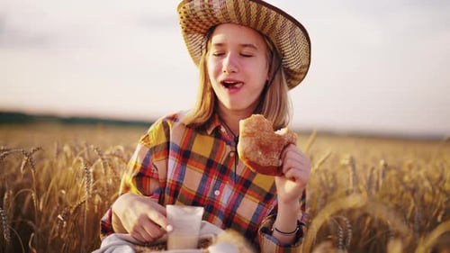 Young Girl Farmer in Sun Hat Eats Loaf of Bread Biting It Drinking Milk Sits on Wheat Field with