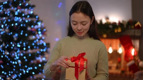 Slim Charming Asian Young Woman Standing in Living Room on Christmas Eve Opening Gift Box with