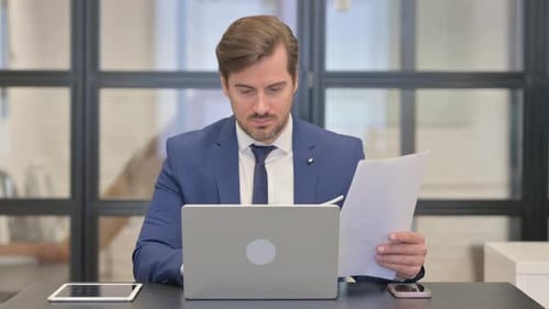 Man in Suit Reading Papers at Computer