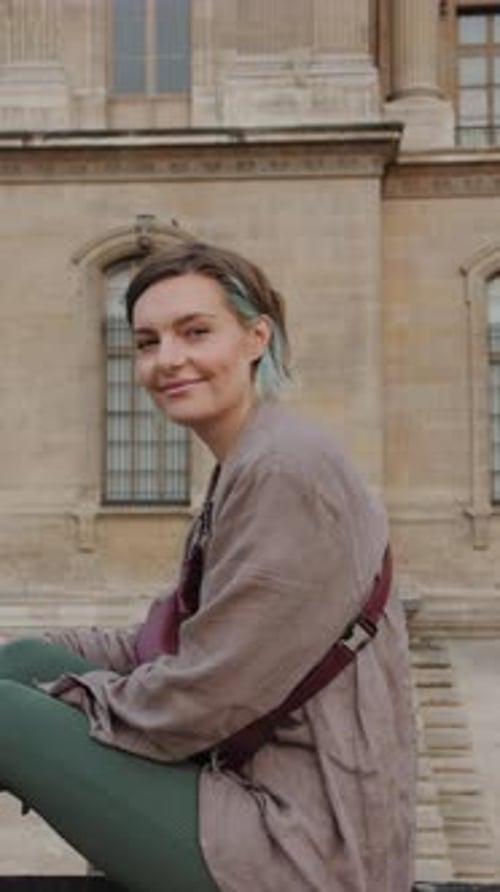 A woman wearing a brown jacket and green pants is sitting on a ledge
