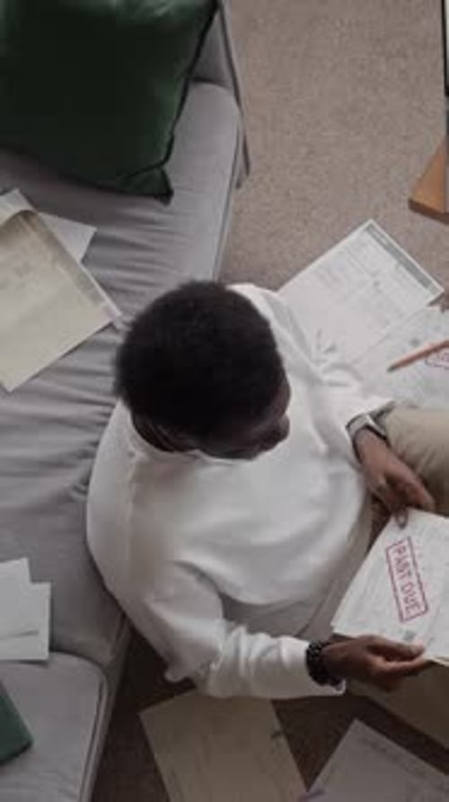 Vertical of Stressed Black Man Surrounded by Overdue Bills Using Laptop