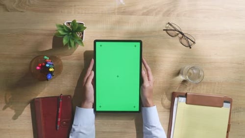 Top down view of man's hands holding a green screen tablet on a wooden desk