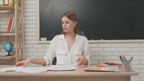 Young Woman College Teacher at Desk in Classroom in Front of Chalkboard Organizing Test Papers of