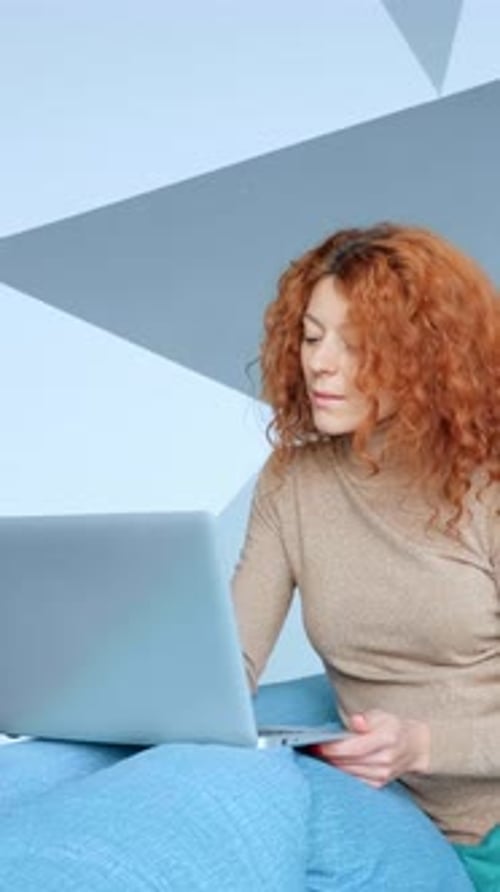 Woman with Curly Red Hair is Focused on Using Laptop While Sitting on Cozy Bed with Colorful