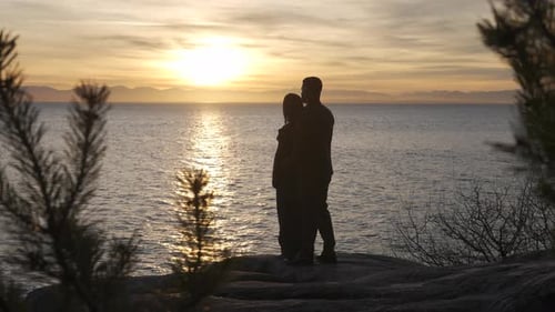 Young couple in love hugging and standing together watching the sunset near the sea on a cloudy day