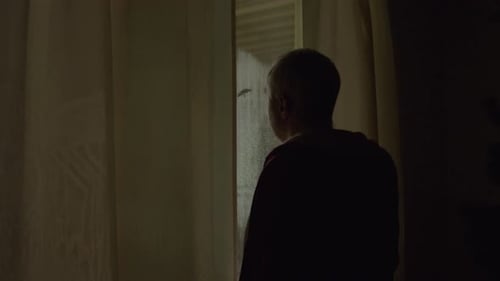 Man Looking Through Window Curtains As Police Sirens Flash At Night