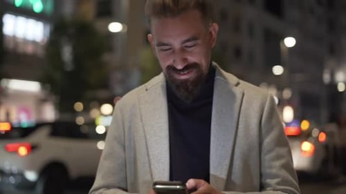 Cheerful Caucasian Man Walking at Night in a Lively City Using Phone to Send Online Messages