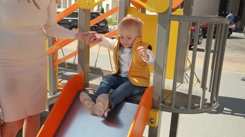 Mother helping her baby walking up the stairs on playground and riding down the slide