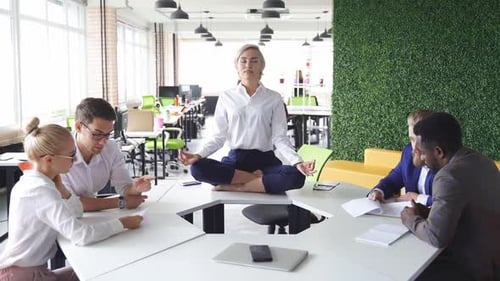 Young Caucasian Woman Meditating in Office Sitting on Table in Lotus Position While Colleagues are