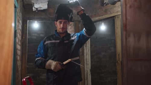 Professional Welder in Safety Suit Puts Up Protective Mask Holding Electrode Holder and Smiling