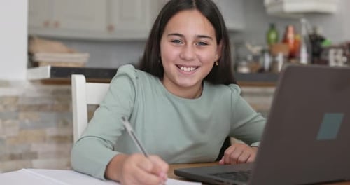 Female child smiling on camera while doing homework using laptop indoor - Back to school