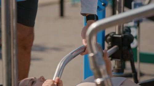 Woman lifting weights, man assisting outdoors, sunny day
