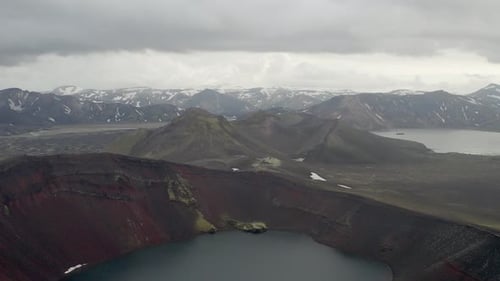 Aerial view over Ljótipollur crater with red rocks and a lake in icelandic highlands - Snowy peak