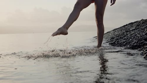 Unrecognizable Sexy Slender Young Woman Enters Water Along Stone Beach