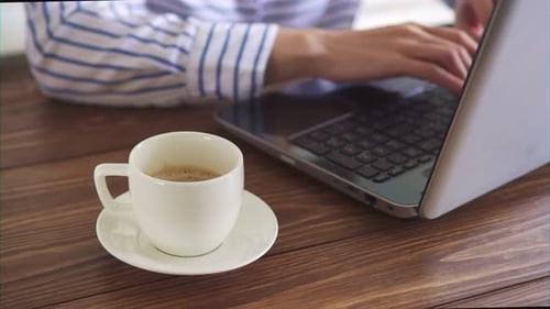 Weiße Kaffeetasse, auf einem hölzernen Arbeitstisch. Hintergrund, ein Mädchen in einem gestreiften Hemd arbeitet an einem Laptop
