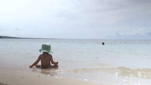 A Small Child in a Children's Panama Sits on the Seashore