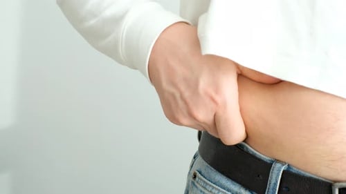 A male hand holding excessive belly fat on gray background, overweight concept.