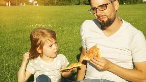 Happy Family Father and Child on Picnic Eat Pizza in Summer Park on Green Lawn at Sunset