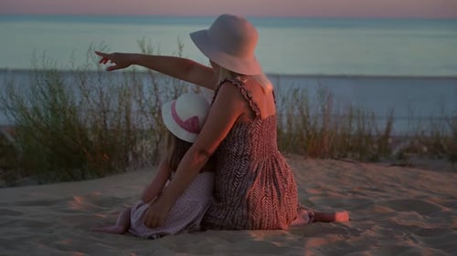 Mother and Daughter Sit on Beach and Look at Blue Sea at Sunset Summer Vacation