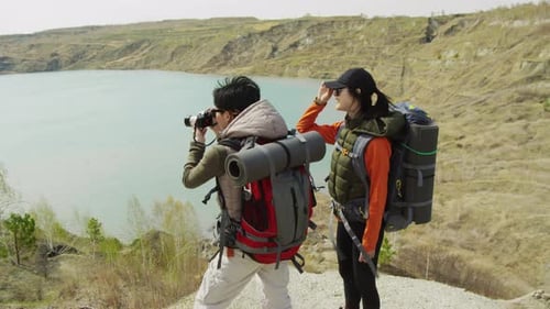 Two Asian Tourists Standing On Rocky Coast Of Lake, One Making Photo With Camera