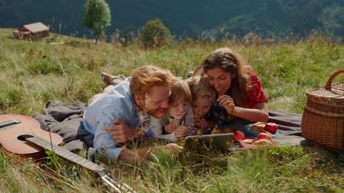 Positive Family Holding Tablet Computer on Picnic Lying Green Grass Mountain Hill
