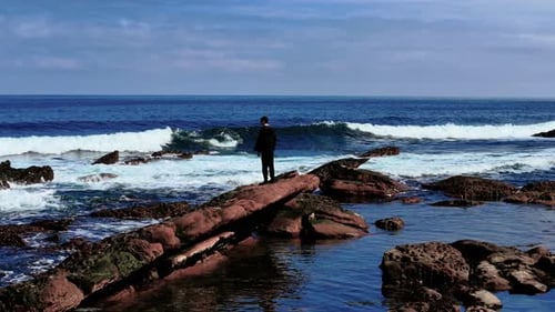 Young Man Standing on Red Rock Looking at Ocean Waves Teenager Observing Crashing Surf From Rocky