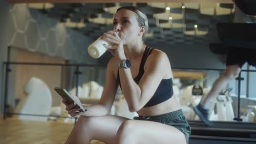 Woman Drinking Water and Using Phone in Gym