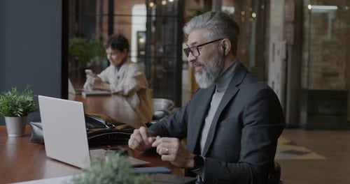 Mature Businessman Making Online Video Call with Laptop and Wireless Earphones Working in Cafe