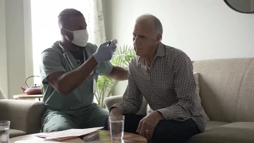 Home Caregiver in Face Mask Checking Temperature of Senior Man on Sofa Adult