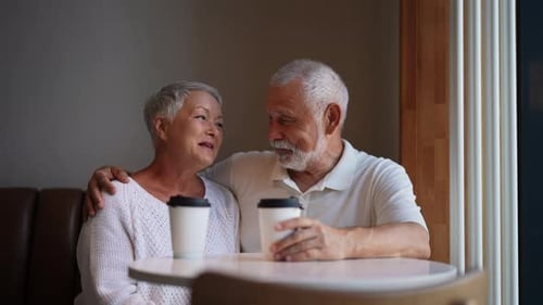Cheerful Elderly Couple Enjoying Coffee and Conversation at Cozy Cafe Table Sitting Hugging By