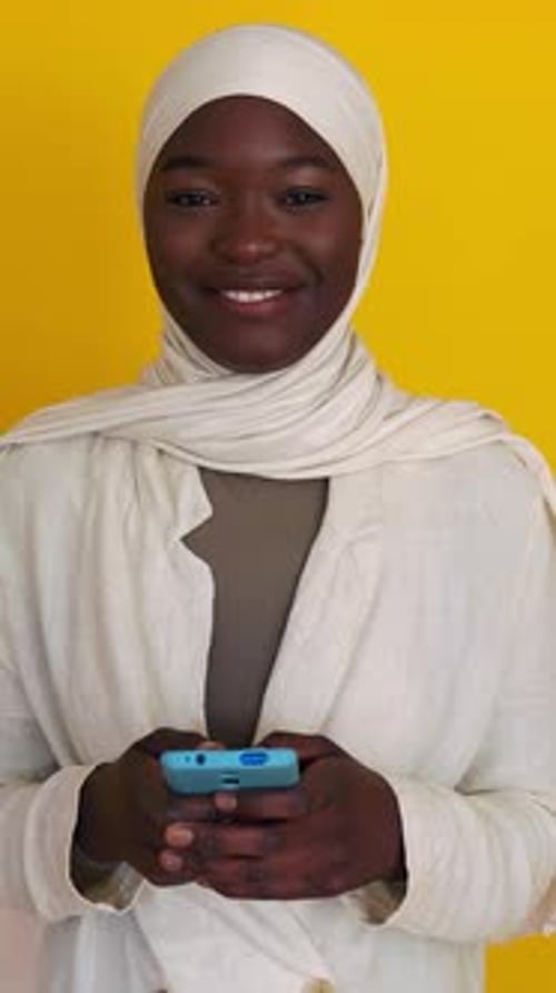African Muslim Woman with Phone Smile at Camera Over Yellow Background