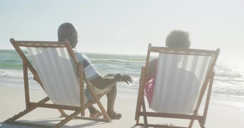 Happy senior african american couple sitting on deck chairs at beach, in slow motion