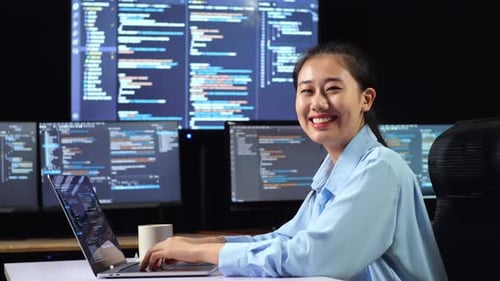 Seitenansicht einer asiatischen Programmiererin, die in die Kamera lächelt, während sie Code auf einem Laptop schreibt