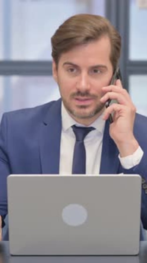 Serious Businessman Talking on Cell Phone While Using Laptop