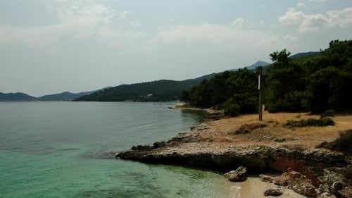 A calm coastline with clear water, rocky shore, green trees, and distant hills under a cloudy sky.