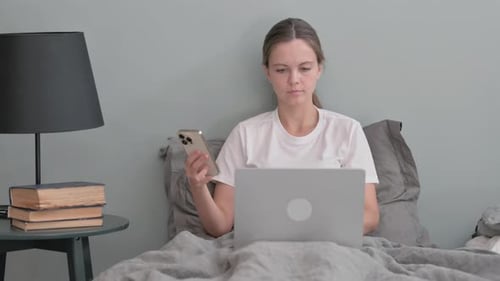 Woman Works on Laptop and Phone in Bed