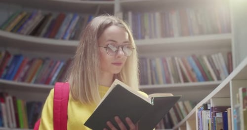 Close Up of Young Caucasian Pretty Blonde Female Student in Glasses Standing in Library and Reading