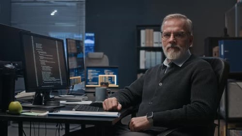 Portrait of Senior Programmer at Desk with Code on Computer Screen at Night