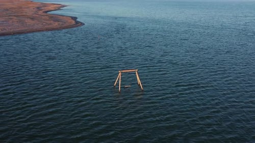 Aerial view above abandoned childhood wooden swing set single chair in ocean sea water by flat brown