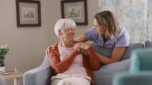 Smiling Woman Embraces Senior Woman Sitting on Couch