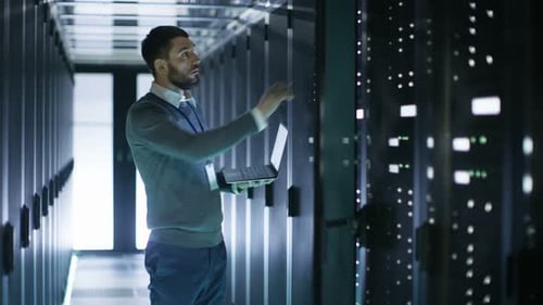 Male IT Technician Working on a Laptop Standing Before Open Server Rack Cabinet in Big data Center.