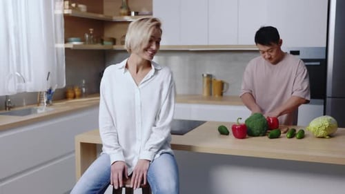 Woman Sitting as Man Prepares Vegetables in Kitchen