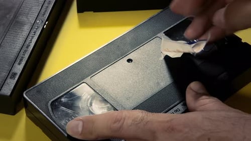 Applying a white label on a VHS tape, with the handwritten text 1990s. Reverse close-up shot (almost