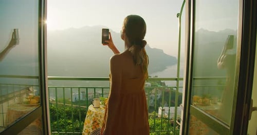 Woman Taking Photo from Sunlit Balcony Overlooking Mountains