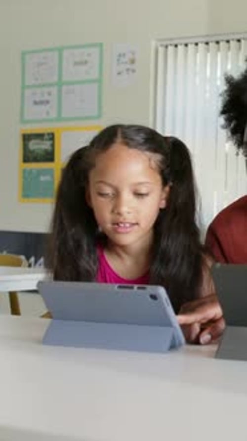 Vertical video: Following adult pointing, girl tapping grey-blue tablet in folio at table, learning