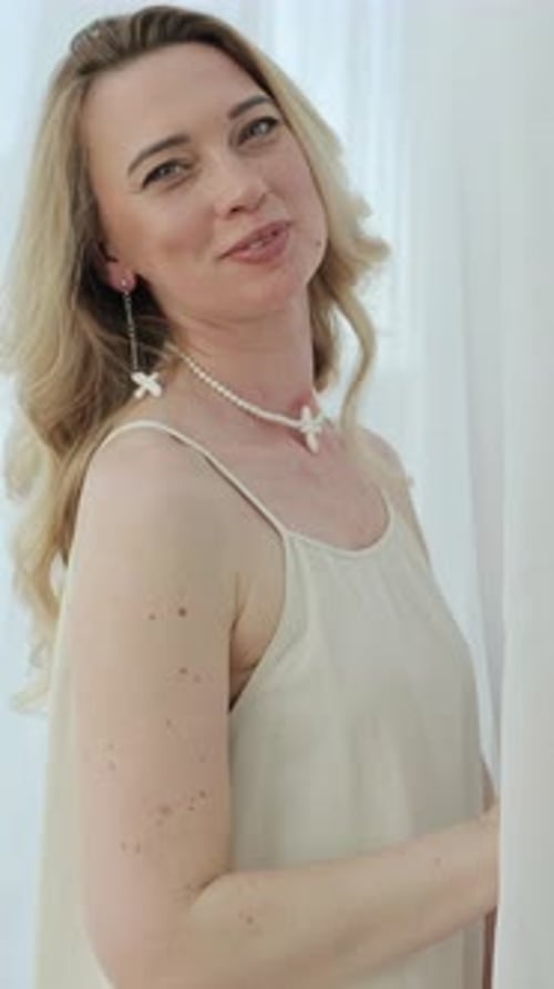Femme blonde souriante se tournant vers la caméra dans une pièce lumineuse avec une énergie calme, paisible et positive