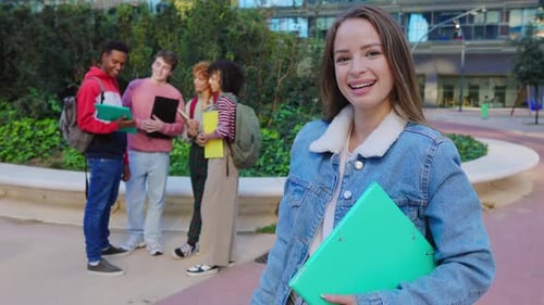 Estudante universitário sorridente segurando um fichário no campus com amigos ao fundo